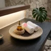 A luxury spa setting featuring a coiled seagrass tray holding rolled white towels and essential oil bottles on a marble counter.