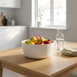 A pristine white lacquered bamboo bowl filled with fresh, vibrant green apples, serving as a modern centerpiece on a light wooden dining table.