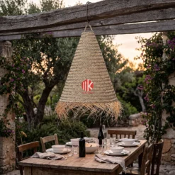 A conical palm leaf lampshade with a fringed edge hangs from a tree branch at an outdoor evening wedding.