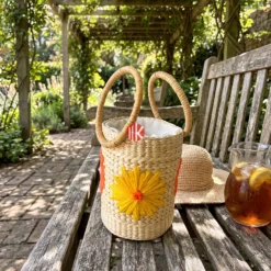 Floral Embroidered Seagrass Round Handbag with Circular Handles 7 Cylindrical woven seagrass bag with circular handles and floral embroidery on a weathered wooden bench in a sunlit garden