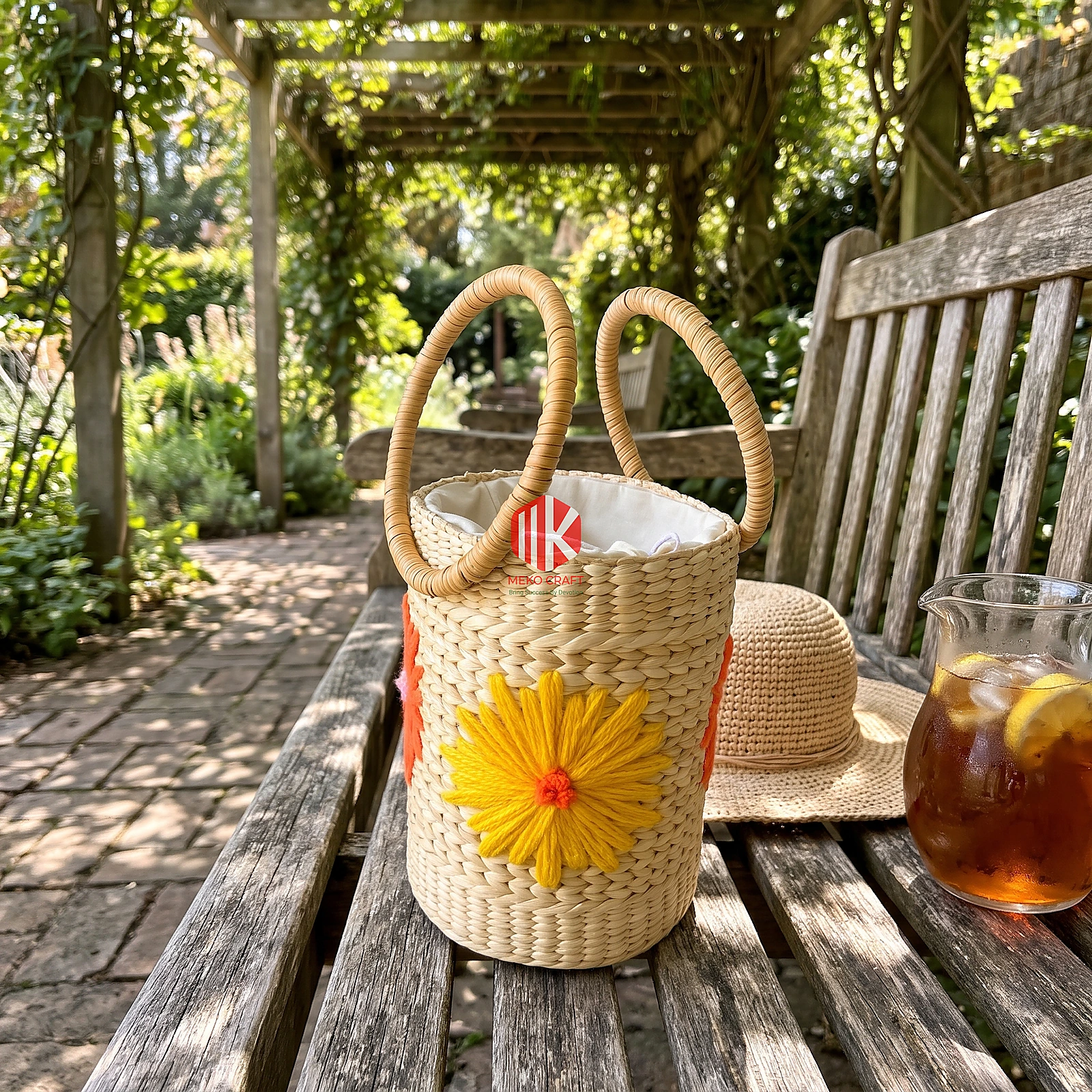 Floral Embroidered Seagrass Round Handbag with Circular Handles 4 Cylindrical woven seagrass bag with circular handles and floral embroidery on a weathered wooden bench in a sunlit garden