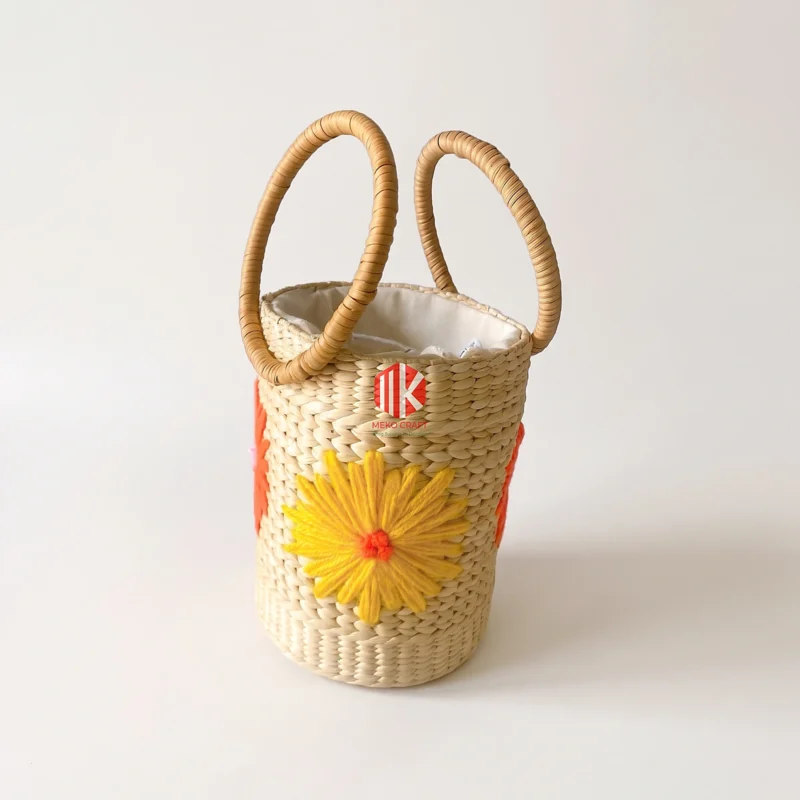 Floral Embroidered Seagrass Round Handbag with Circular Handles 10 A studio shot of a cylindrical seagrass handbag with yellow floral embroidery and circular handles on a pure white background.