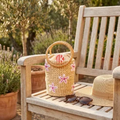 A floral water hyacinth handbag resting on a sun-drenched wooden table at a coastal resort, next to a pair of sunglasses.