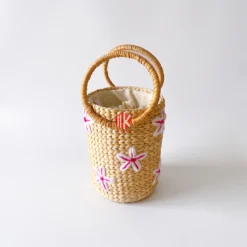 Studio shot of a cylindrical water hyacinth handbag with white and pink floral embroidery and two round top handles on a white background.