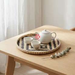 A round patterned bamboo tray rests on a light oak coffee table holding a ceramic teapot and two small tea bowls.