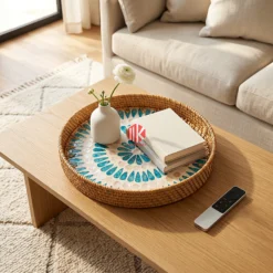 A patterned rattan and mother of pearl round serving tray rests on a light-colored console table, holding a small vase and a book.