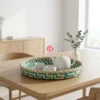 A round bamboo tray with a teal herringbone pattern sits on a light oak dining table holding a white ceramic teapot and two cups.