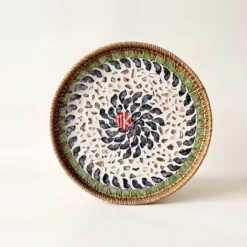 Detailed close-up of a circular mother of pearl serving tray displaying a black shell swirl inlay, green border tiles, and handwoven rattan rim
